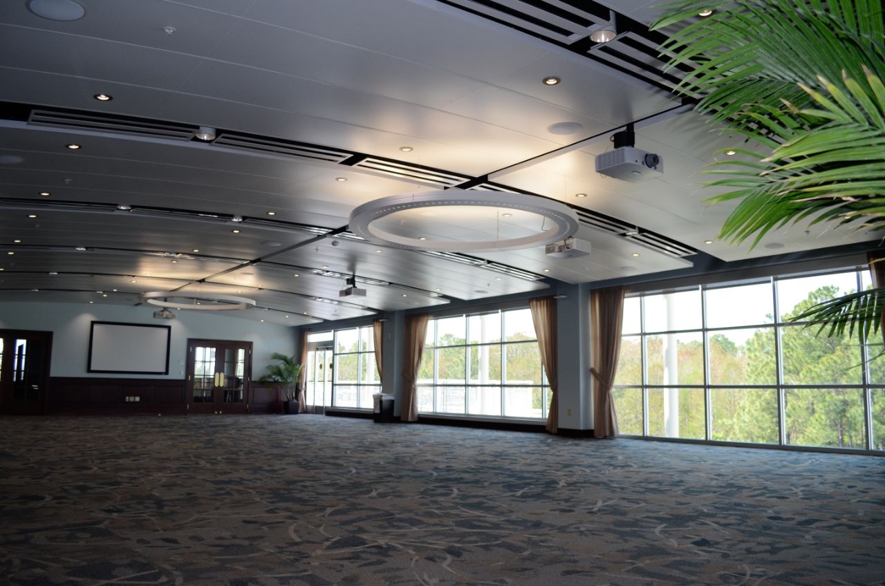 Large open room with floor to ceiling windows and blue patterned carpet has large circular light fixtures hanging from the ceiling as well as a projection screen on the far end of the room.