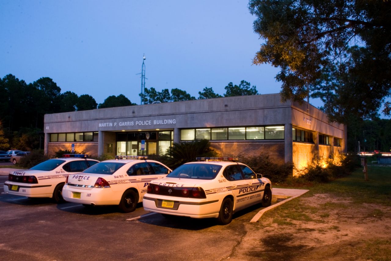 Three white police cars are parked in front of a brick building with white trim; Building windows are lit from within at dusk