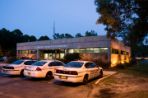 Three white police cars are parked in front of a brick building with white trim; Building windows are lit from within at dusk