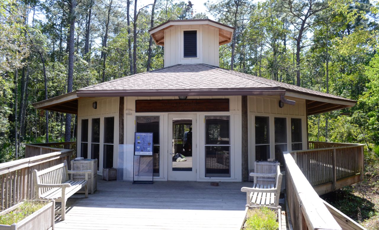 One story off-white hexagonal building with wood paneling and windows on all sides sits on a wide wooden walkway that leads into UNF’s wooded natural preserve.