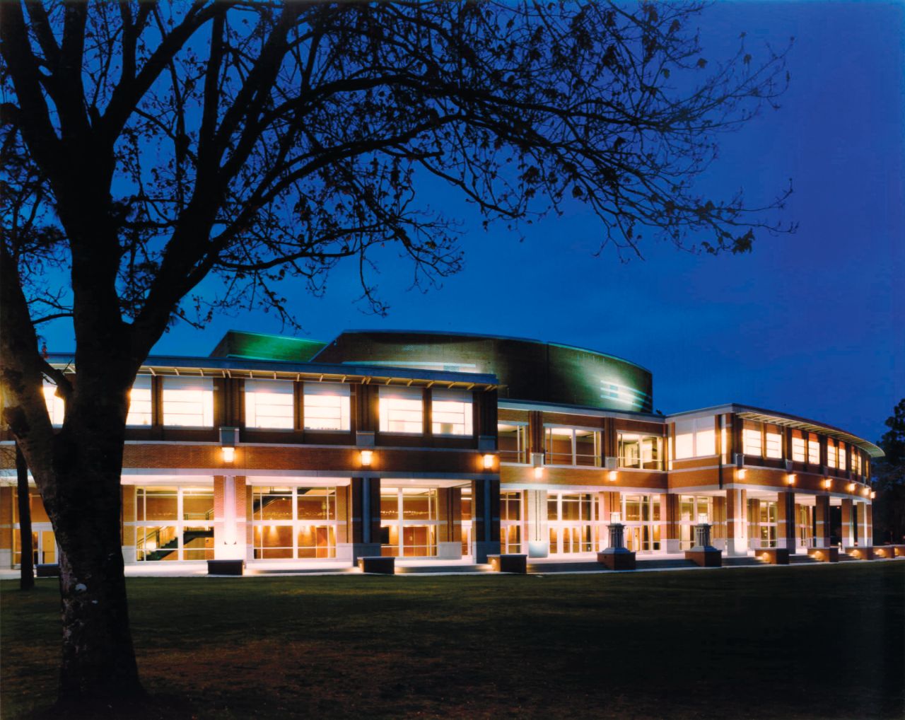 In a late evening photo, the curved façade of a brick building with large windows and yellow spotlights suspended above columns that are spaced across the front; the building sits on a grassy lawn with a shadowy tree in front.