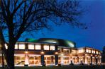 In a late evening photo, the curved façade of a brick building with large windows and yellow spotlights suspended above columns that are spaced across the front; the building sits on a grassy lawn with a shadowy tree in front.