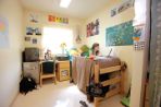 Caucausian student studies with a textbook and computer on an elevated wood single bed that is positioned next to a small wooden desk inside a white-walled room decorated with colorful posters.