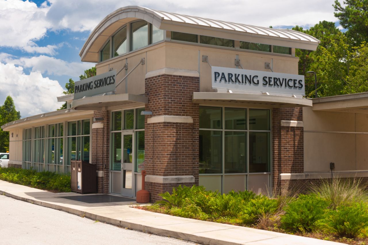 One-story red brick building with off-white trim and an arched metal roof sits on a curb with shrubbery in front.