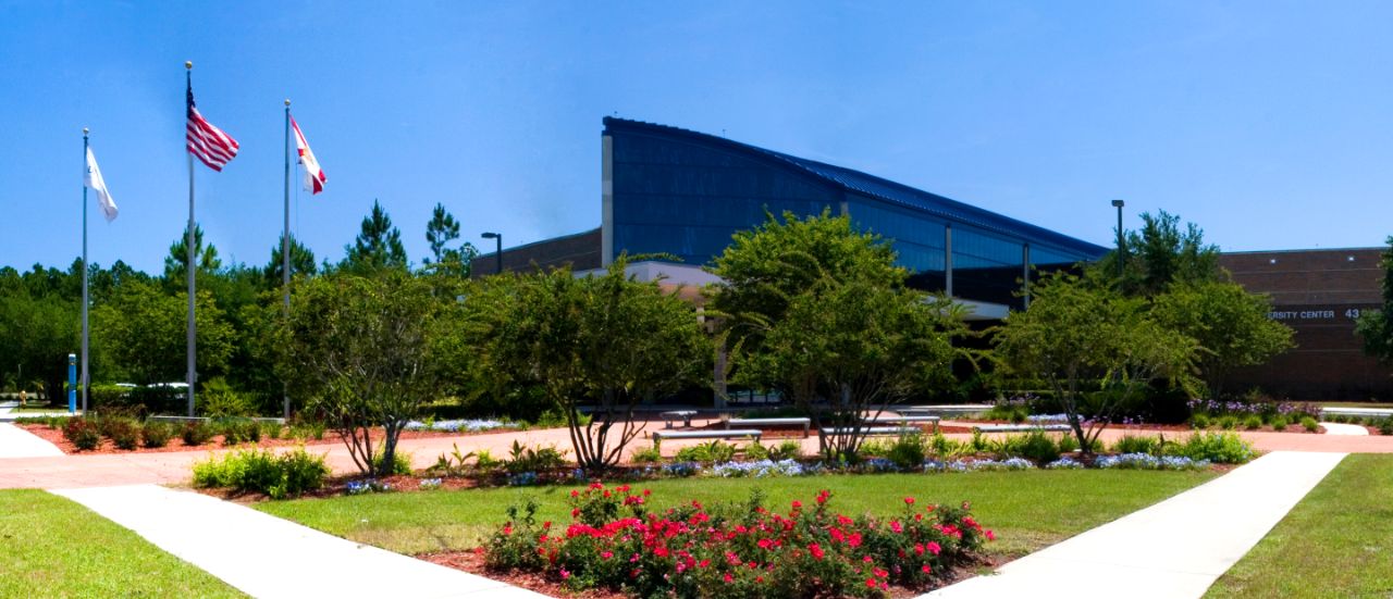 Dark blue building with arching mirrored windows with red flowers and trees in the foreground between sidewalks approaching the building