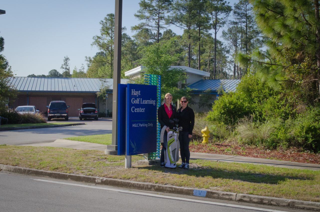 Two female students wearing dark athletic pants and jackets stand next to a blue and green sign that says Hayt Learning Center; One of the students holds a green and gray golf bag; In the background, behind a cluster of shrubbery is a one story brick building with a metal roof.