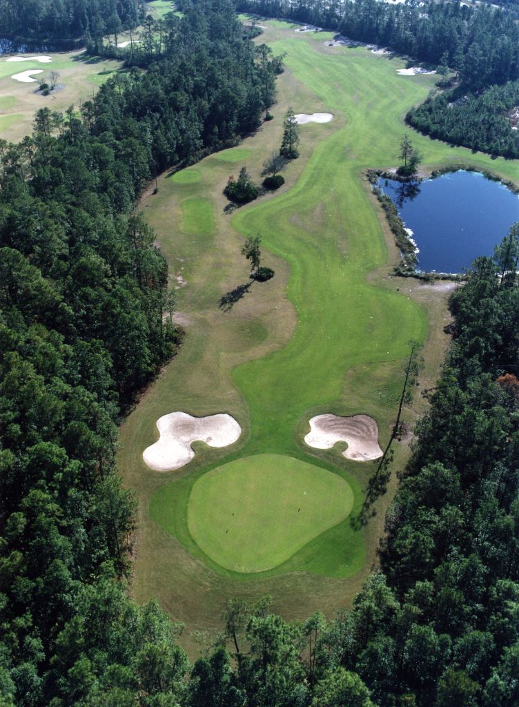 Aerial view of golf course with two sand traps near a putting area and a lake in the distance, surrounded by dense trees.