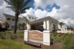 White and brown sign with the words Osprey Fountain sits in a landscaped plant bed in front of a large white stucco building with a covered driveway