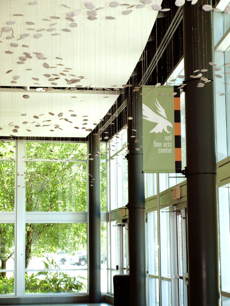 Small white disks are suspended by thin metal wire along the length of a window-lined corridor; a vertical banner hanging from a black column on the right depicts a white osprey emblem and the words “UNF Fine Arts Center.”