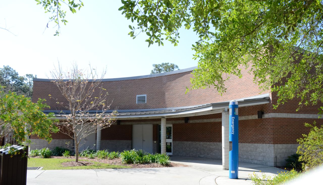 Small one-story brick building with white trim and white columns; a short metal awning arches across the front of the building which opens out to a landscaped courtyard