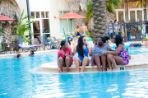 Four African American female students wearing shorts, tank tops and sunglasses talk while sitting on the edge of white pool deck with their feet in blue water in the pool