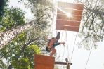 From below, a student wearing a harness leaps between two suspended wooden platforms; in the background, a group of other students wearing harnesses wait on a structure to cross the suspended platforms.