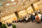 A crowded room of spectators sitting in chairs gathered around circular tables watches and listens to a speaker presenting at a podium.