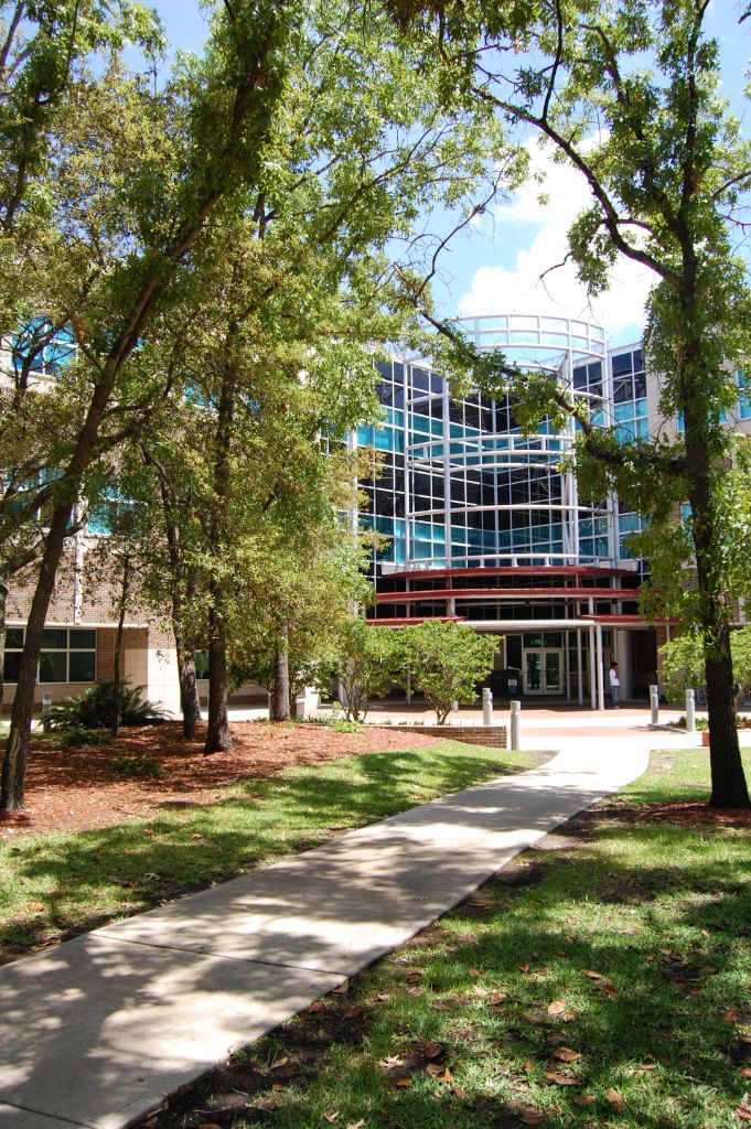 Three-story steel and glass building with curved metal awning across the front; a sidewalk leading up to the front entrance of the building is shaded by large trees