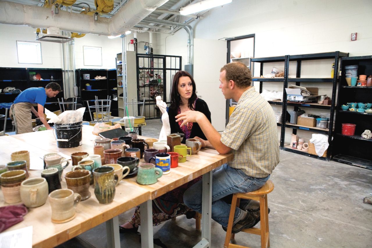 Caucasian female student with brown hair wearing black shirt talks to Caucasian male professor with brown hair wearing beige shirt; They are sitting at a table gesturing to a small white sculpture; Also on the table are several ceramic bowls and vases in various bright colors.