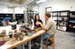 Caucasian female student with brown hair wearing black shirt talks to Caucasian male professor with brown hair wearing beige shirt; They are sitting at a table gesturing to a small white sculpture; Also on the table are several ceramic bowls and vases in various bright colors.