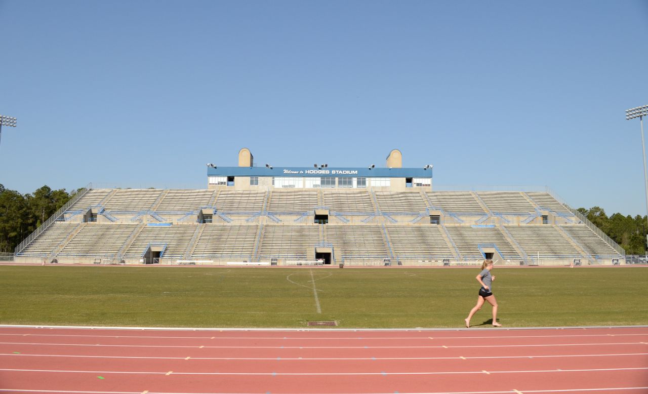 A girl wearing shorts and a t-shirt runs along the red track that surrounds a large grass field; on the far side of the track is a two-level cement stadium with metal bleachers and blue railings; atop the stadium is a wide press box with tall windows and the words Welcome to Hodges Stadium on a sign in front.