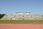 A girl wearing shorts and a t-shirt runs along the red track that surrounds a large grass field; on the far side of the track is a two-level cement stadium with metal bleachers and blue railings; atop the stadium is a wide press box with tall windows and the words Welcome to Hodges Stadium on a sign in front.