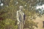 Bronze statue of Martin Luther King, Jr. standing upright and holding a book; Statue sits on engraved granite pedestal in a flower bed with trees and a building in the background.