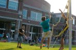 Three students play volleyball on a grassy lawn; One student in a teal polo shirt is jumping up to spike the ball while another student in a black shirt is trying to block the hit on the other side of the net. In the background, dozens of students mill around, gathered in groups on the lawn or sitting on the steps of a two-story brick building in the background.