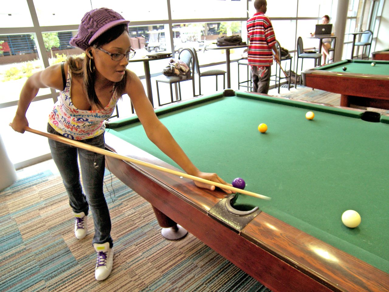 A female African American student wearing jeans, a tank top and a purple knit hat leans over a pool table to shoot a ball in an area of the game room with other pool tables in the background that sit along a window-lined wall.