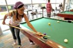 A female African American student wearing jeans, a tank top and a purple knit hat leans over a pool table to shoot a ball in an area of the game room with other pool tables in the background that sit along a window-lined wall.