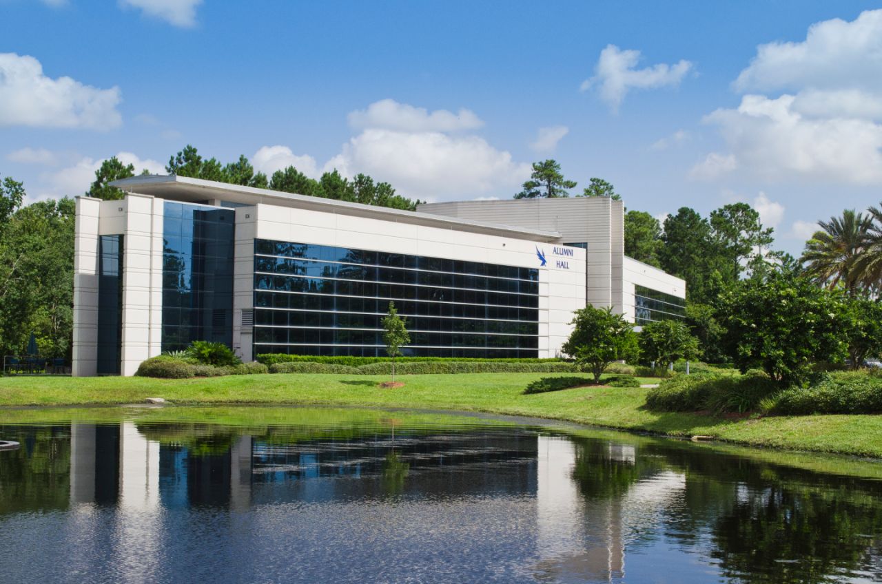 A three-story glass and stucco building is lined by neatly trimmed shrubs on a large grassy lawn with trees that sits on a pond.