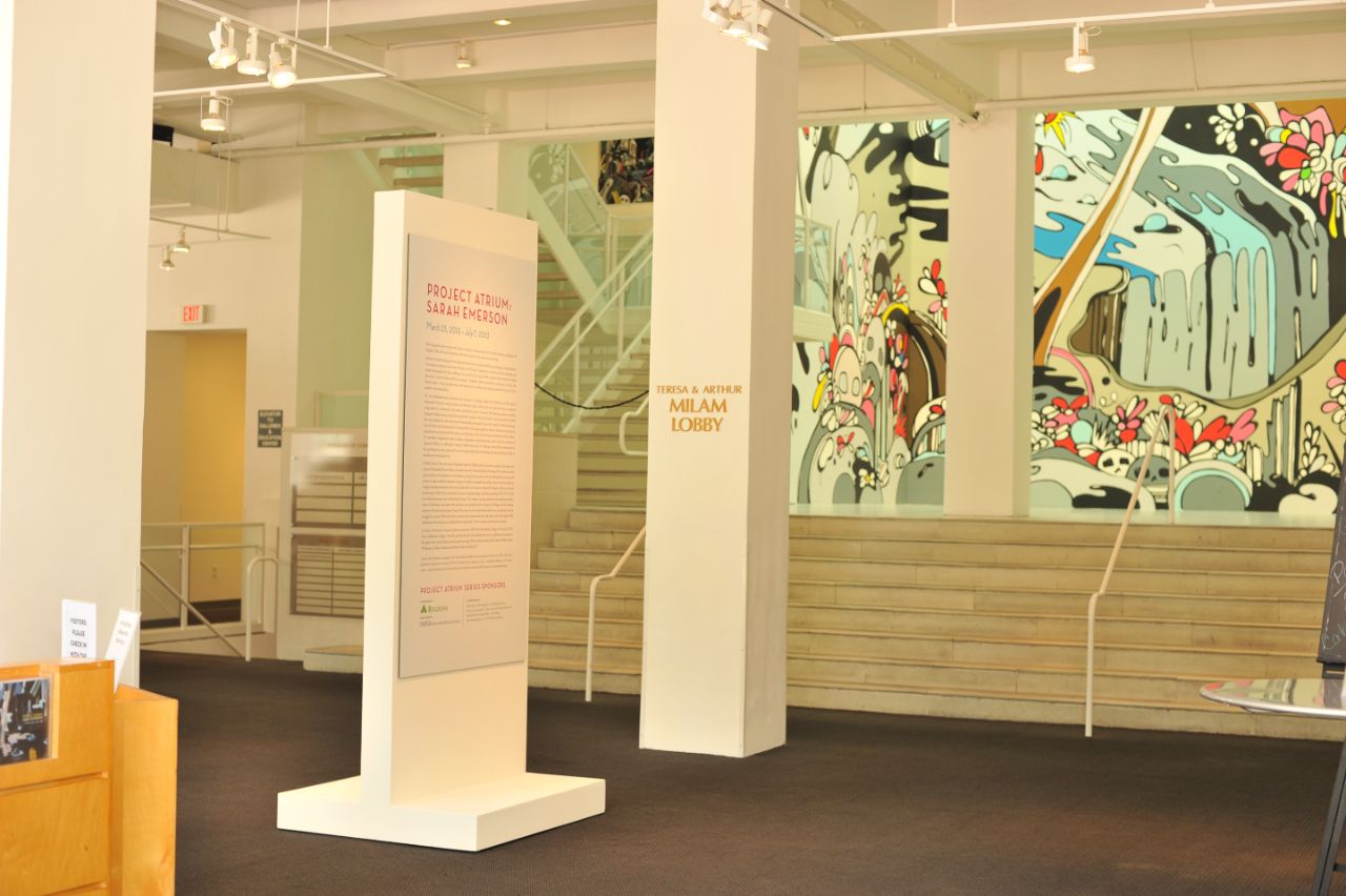 Open lobby of the museum with concrete floors and white walls with a short stair case leading up to a landing space with a bright mural painted on the back wall; white square columns split the lobby and a tall white directional sign sits in the middle of the space.