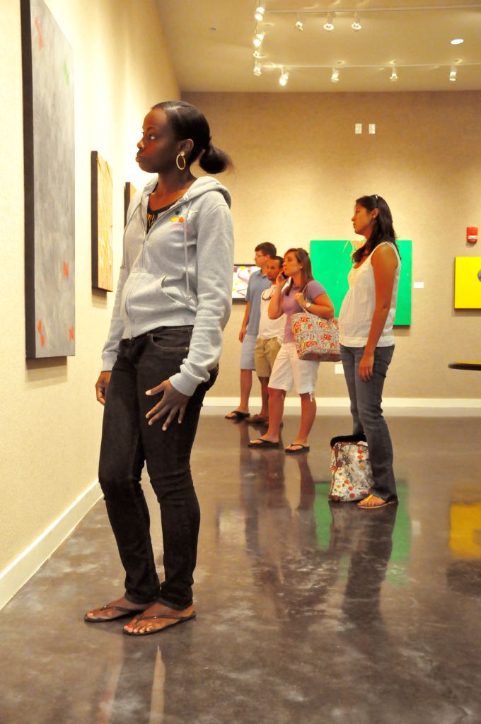 Five male and female students wearing jeans and t-shirts are standing in a room with a gray concrete floor and off-white walls where they are looking at several colorful paintings on canvas.