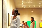 Five male and female students wearing jeans and t-shirts are standing in a room with a gray concrete floor and off-white walls where they are looking at several colorful paintings on canvas.