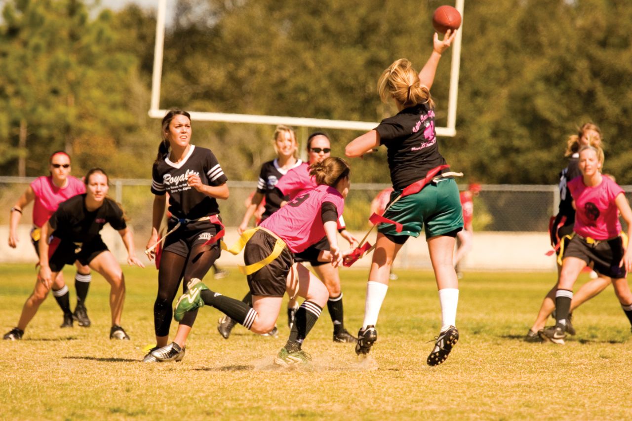 Two teams of female students — one wearing pink shirts and one wearing black shirts — stand ready on a grass field as a female student throws a football downfield and an opposing team member attempts to block her; a white football goal post stands in the background