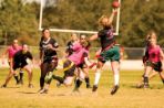 Two teams of female students — one wearing pink shirts and one wearing black shirts — stand ready on a grass field as a female student throws a football downfield and an opposing team member attempts to block her; a white football goal post stands in the background