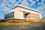 A three-story brick, stucco and glass building sits on a landscaped lawn along a paved road.