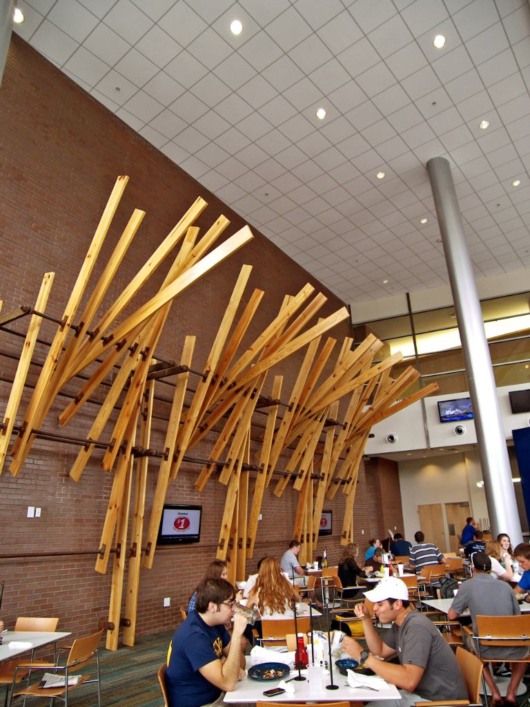 Students eat grill-style food at white square tables in a two-story room that has a tall metal support pole toward the back and a sculpture of wide wooden panels protruding from an exposed brick wall.