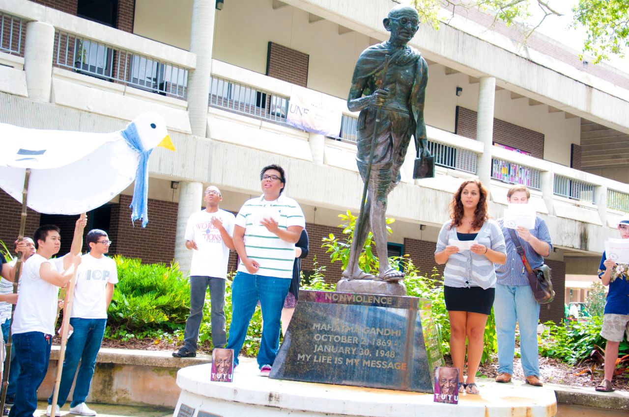 Students standing on the edges of plant beds and the wide base of a bronze Mahatma Gandhi statue gesture together as they sing a song; To the left a small group of students hold a large kite depicting a peace dove