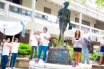 Students standing on the edges of plant beds and the wide base of a bronze Mahatma Gandhi statue gesture together as they sing a song; To the left a small group of students hold a large kite depicting a peace dove