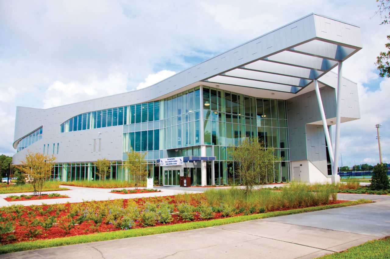 Three-story glass and steel building with a large steel awning sits on a landscaped pathway edged with plant beds and trees.