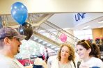 A father wearing a baseball hat smiles and shows his daughter and wife a teddy bear wearing a UNF shirt while his daughter holds a blue t-shirt with North Florida Ospreys printed on the front; they are in the central lobby of the bookstore with clothing racks and balloons in the foreground and stairs in the background.