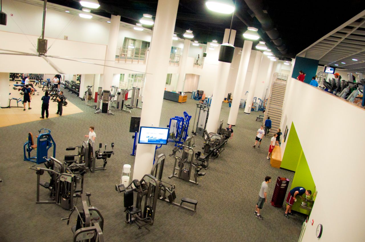 Large indoor open area has two-story ceilings and contains rows of various weight and exercise machines where students are milling around.