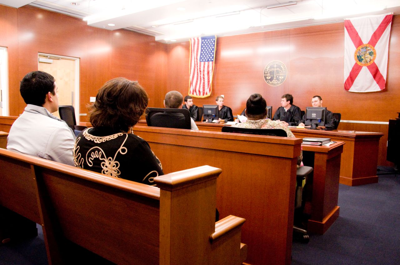 In a wood-paneled room, several male students wearing black judicial robes sit at the front of a room where they face representatives at two desks facing them. In benches behind the desks are more student spectators watching the proceedings.