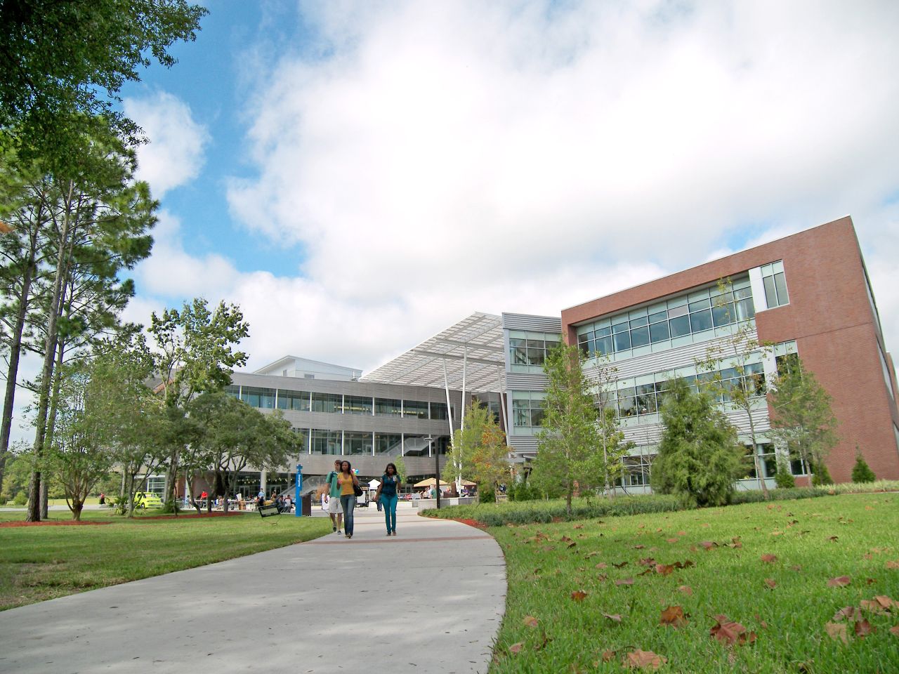 Two side-by-side three-story brick, steel and glass buildings with a slanted metal awning covering the walkway between them; in the foreground, a wide sidewalk winds through a grassy lawn approaching the buildings where several students walk together.