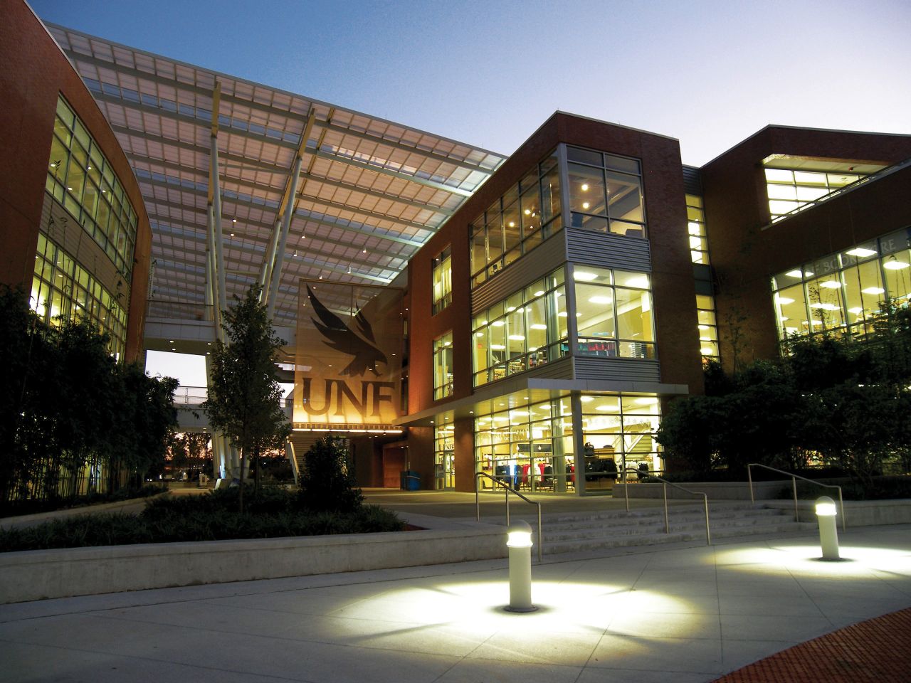 Two side-by-side three-story brick, steel and glass buildings with a slanted metal awning covering the walkway between them; suspended in the walkway is a two-story mesh banner featuring the UNF logo; in the foreground are two light fixtures illuminating a wide sidewalk in front of the buildings.