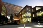 Two side-by-side three-story brick, steel and glass buildings with a slanted metal awning covering the walkway between them; suspended in the walkway is a two-story mesh banner featuring the UNF logo; in the foreground are two light fixtures illuminating a wide sidewalk in front of the buildings.