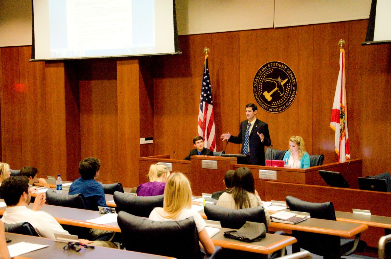 In a wood-paneled room, a male student wearing a suit and tie stands up at a podium in front of an official Student Government Senate seal, addressing a group of student senators, each sitting in a leather chair at a desk facing the podium.