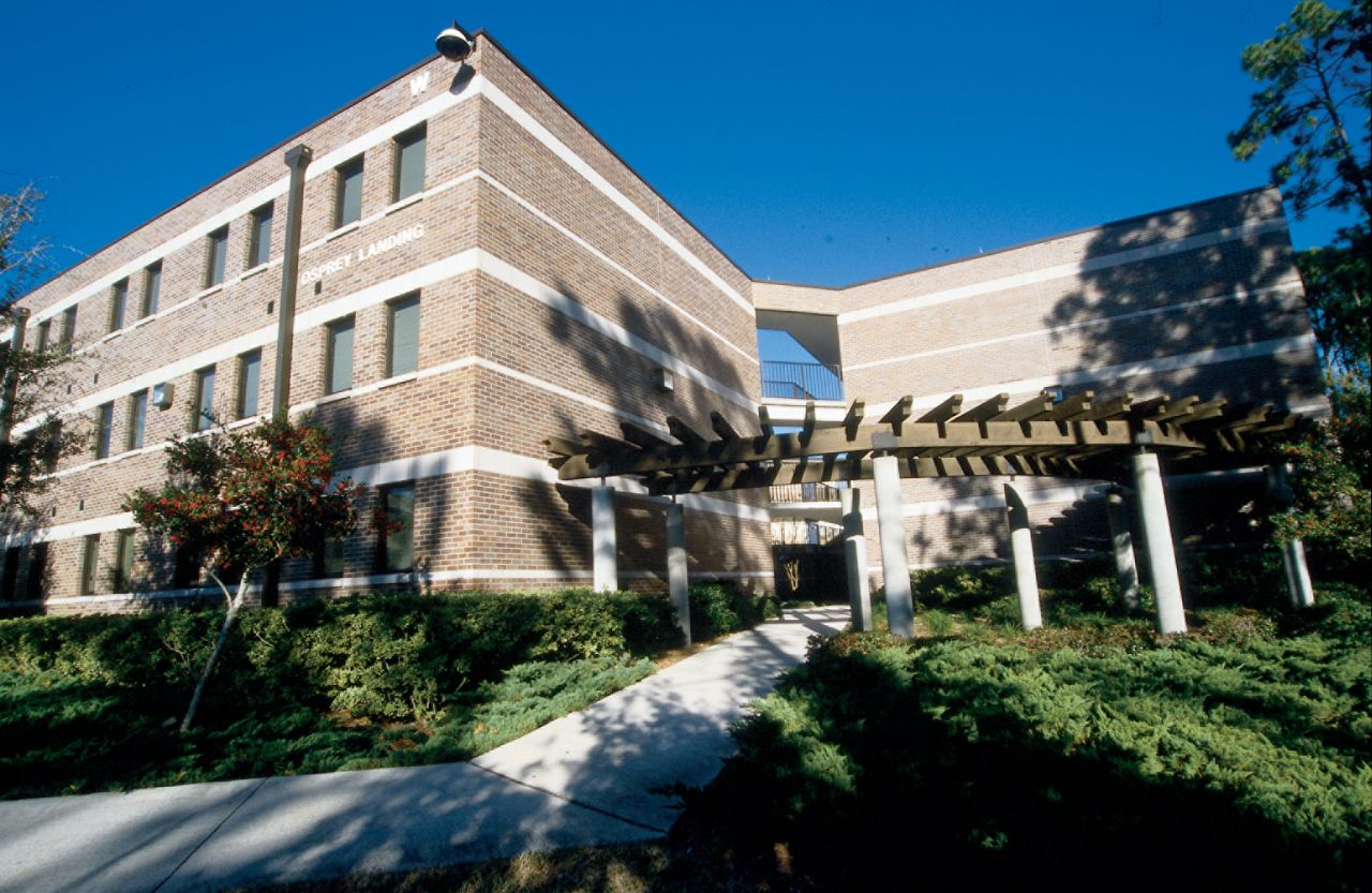 Three-story brick building with a corner entrance covered by a wooden trellis with white columns sitting behind a landscaped path flanked by shrubbery.