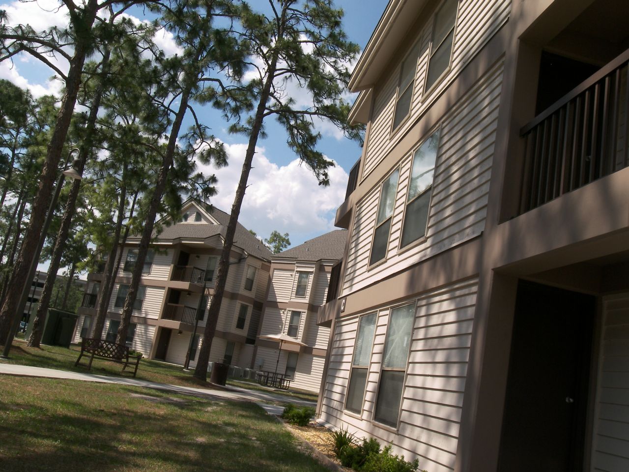 Two three-story buildings with off-white paneling and brown trim sit on a grassy area criss-crossed with concrete sidewalks; benches and picnic tables are situated along the pathways near the buildings and several tall pine trees are scattered in the area