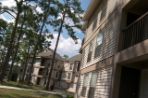 Two three-story buildings with off-white paneling and brown trim sit on a grassy area criss-crossed with concrete sidewalks; benches and picnic tables are situated along the pathways near the buildings and several tall pine trees are scattered in the area