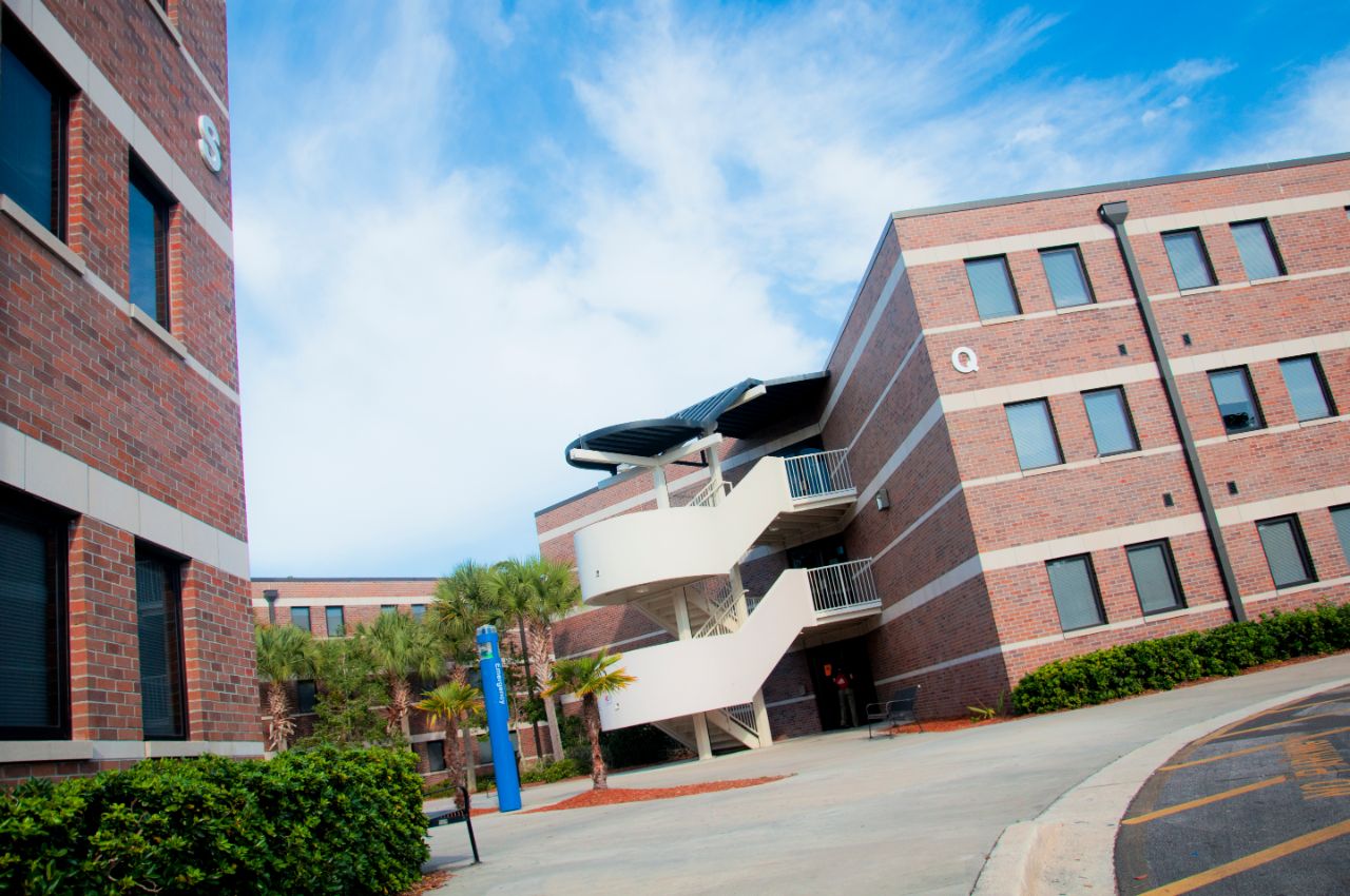 A cluster of three red brick three-story buildings with white trim surround a concrete courtyard that is broken up by landscaped plant beds around perimeter and in the middle that contain shrubs and palm trees. Metal benches are positioned near a few of the plant beds.