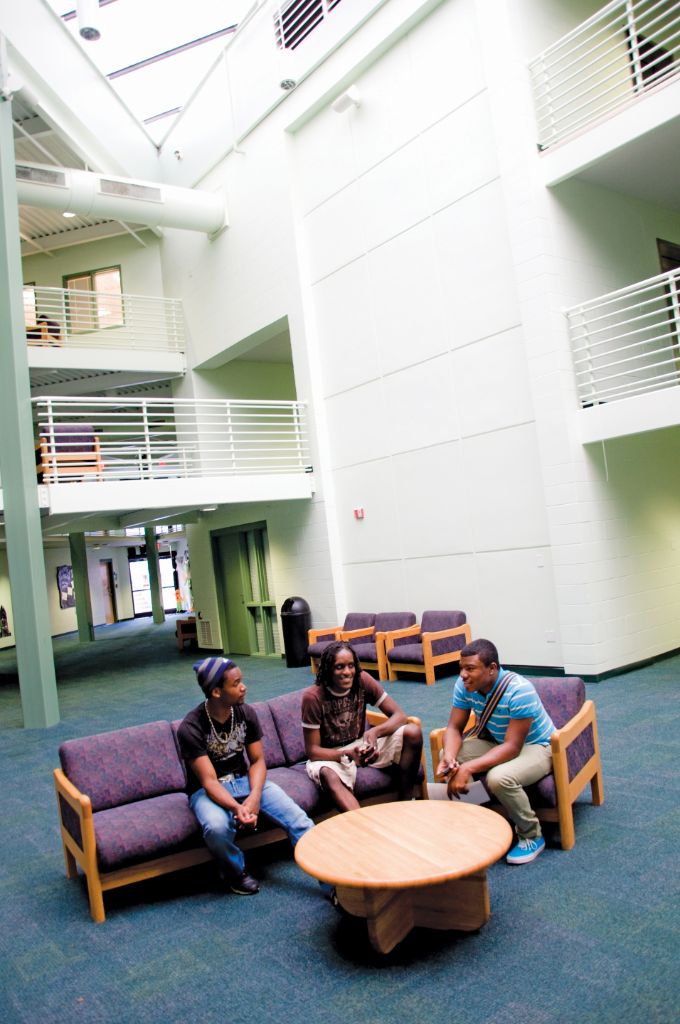 A male and female student sit on a purple couch and talk to a male student sitting on a purple chair next to them; the furniture is positioned around a round wooden coffee table in a large, open white room with blue carpet and several balconies with white railings.