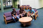 A male and female student sit on a purple couch and talk to a male student sitting on a purple chair next to them; the furniture is positioned around a round wooden coffee table in a large, open white room with blue carpet and several balconies with white railings.